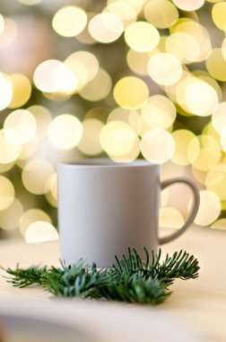 Yeni yıl tebrik kartı. Ahşap masada Noel ağacı dallarıyla süslenmiş gri bir fincan sıcak çikolata var. Bulanık sarı ışıklar arka planda. Kutlama içkileri. Uzayı kopyala Doku