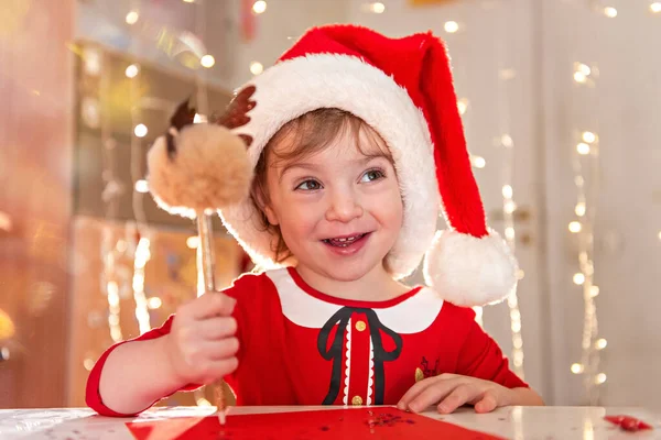 Kırmızı elbiseli ve şapkalı küçük mutlu kız Noel Baba 'ya mektup yazıyor. Çocuk Noel için bir dilek diler. Bebek gülümser, kağıda kalemle yazar. Mesaj için yer, kart. Boşluğu kopyala