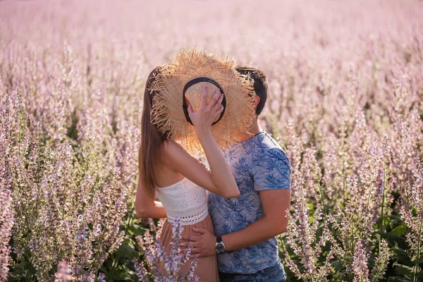 一对恋爱中的快乐夫妻躲在草帽后面 在夕阳西下的一片盛开的圣园前接吻 一个年轻人热情地拥抱 牵着一个漂亮姑娘的手 情侣周末旅行 复制空间 — 图库照片