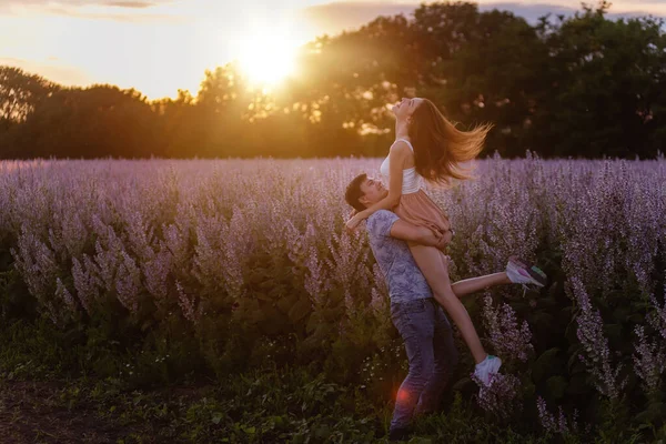 年轻的男人抱着美丽的女人 在夕阳西下的光芒中 旋转着她绕着盛开的紫罗兰花园地 这个女孩的头发向不同的方向飘扬着 一个结婚的提议 复制空间 — 图库照片