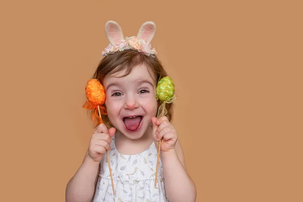 Positive portrait of little girl with rabbit ears on her head, showing her tongue holding an orange green egg. Isolated light orange background, copy space, postcards. Festive child in costume bunny