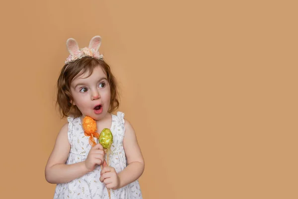 Positive portrait of little girl with rabbit ears on head, looks in surprise, holding an orange green egg in her hands. Isolated light orange background, copy space postcards. Festive child in costume