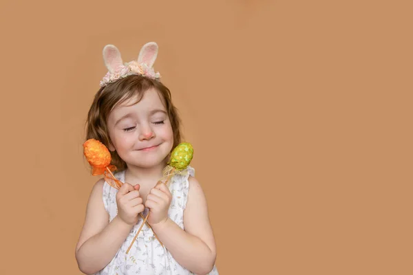 Positive portrait of little girl with rabbit ears on her head, dreams of chocolate, in her hands an orange green egg. Isolated light orange background, copy space, postcards. Festive child in costume