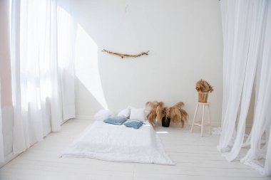 İskandinav iç tarzında minimalist beyaz çatı katı. Ahşap zeminde yatak, pencerelerde hafif havadar tüller vazolarda pampa otları, duvarda tahta dallar var. Modern ev tasarımı