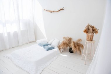 İskandinav iç tarzında minimalist beyaz çatı katı. Ahşap zeminde yatak, pencerelerde hafif havadar tüller vazolarda pampa otları, duvarda tahta dallar var. Modern ev tasarımı