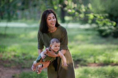 Genç kahverengi saçlı anne, parkta, ağaçların arasında yeşil çimenler arasında bebeğini kollarında sallıyor. Küçük kızı kollarına alan, oynayan, gülen çocuğa sarılan bir kadın. Mutlu bir çocukluk. Yaşam biçimi kopyalama alanı