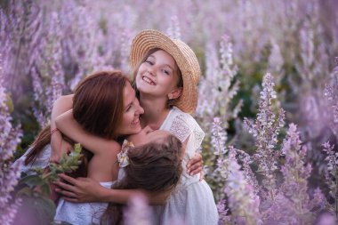 Genç anne mor çiçekli adaçayı tarlasında iki kızını kucaklıyor. Genç kız, bir kadını sevgiyle öper. Saman şapkalı, beyazlar içinde. Çocuklarla seyahat etmek, anne bakımı. Portreyi kapat