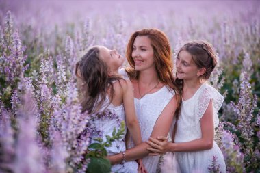 Genç anne mor çiçekli adaçayı tarlasında iki kızını kucaklıyor. Kızlar birbirine sarılır, güler, eğlenir. Saman şapkalı, beyazlar içinde. Çocuklarla seyahat etmek, anne bakımı. Portreyi kapat