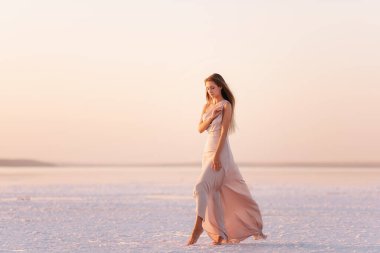 Akşamları havadar, pastel pembeli, pudralı elbisesi olan genç sarışın kadın beyaz kristalize tuz üzerinde yalınayak duruyor. Doğal makyajlı bir kız, saçı gelişiyor. Tuz madenciliği gezisi, gün batımında suda yürümek.