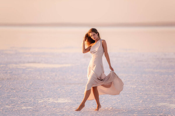 Young blonde woman in an evening airy pastel pink, powdery dress stands barefoot on white crystallized salt. Girl with natural make-up, hair is developing. Salt mining trip, walking on water at sunset