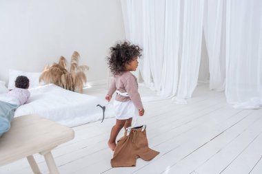 Küçük kıvırcık saçlı Afrikalı Amerikalı kız oynuyor, beyaz yatağın yanında koşuyor, eğleniyor, gülüyor, gülümsüyor. Çok ırklı bir aileyi benimseme kavramı. İskandinav minimalist iç mimarisi, pampa otları. Çocukluk