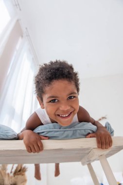 Küçük Afrikalı Amerikalı çocuk eğleniyor, beyaz tahta sandalyeye uzanıyor, ayaklarıyla bocalayıp duruyor. Minimalist iç mekan, otlar, duvarda sürüklenen odun, pencerelerde hava kapıları. İskandinav tarzı.