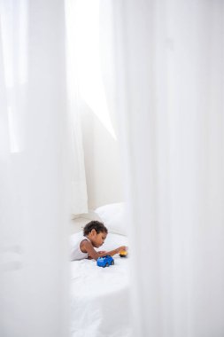 Küçük Afrikalı Amerikalı çocuk tahta zeminde beyaz bir yatakta oyuncak arabalarla oynuyor. Minimalist iç mekan, otlar, duvarda sürüklenen odun, pencerelerde hava kapıları. İskandinav tarzı. Boşluğu kopyala