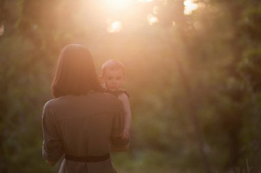 Genç anne, gün batımında kucağında bebeğini kollarıyla parkta tutuyor. Kız arkadan dışarı bakıyor. Kadın kızını öpüyor, sarılıyor. Anne bakımı, velayet. Evlat edinme konsepti. Açık havada yürür