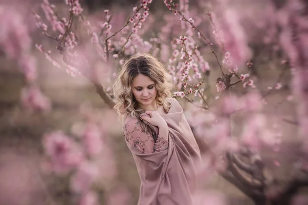 Gentle portrait Young curly blonde woman in brown pleated skirt pink blouse covered shoulders with stole stands in blooming peach gardens at sunset. Girl enjoying walk outside the city, natural beauty