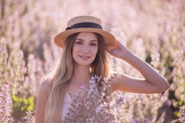 Çiçek açan pembe adaçayı tarlasının arka planında hasır şapkalı genç sarışın kadın. Elinde buket çiçekleri tutan güzel bir kız portresi. Hafta sonu şehir dışında yürüyüş. Tarım dokusu