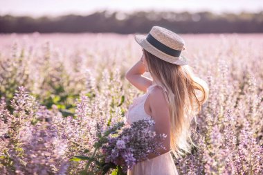 Hasır şapkalı genç sarışın kadın çiçek açan pembe adaçayının arka planına bakıyor. Elinde çiçek tutan güzel bir kızın yakın plan portresi. Tarım dokusu
