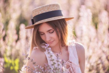 Hasır şapkalı genç sarışın kadın çiçek açan pembe adaçayının arka planına bakıyor. Elinde çiçek tutan güzel bir kızın yakın plan portresi. Tarım dokusu