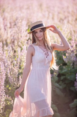Pembe adaçayının çiçek açmış arka planında açık şifonlu elbisesi olan genç sarışın kadın. Hasır şapka tutan güzel bir kızın portresi. Hafta sonu şehir dışında yürüyüş. Tarım dokusu