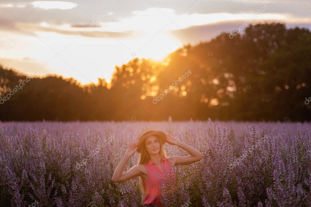 Portrait of a young woman in a magenta shirt, denim shorts, a straw hat on her head against the background of a blooming pink field of sage. Long blond hair in the sun at sunset. Travel out of town