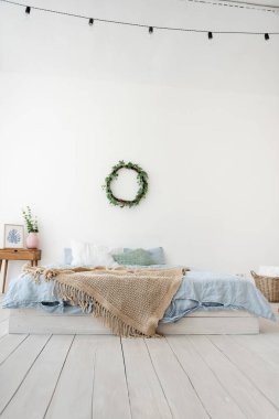 Şık, modaya uygun bir iç mekan İskandinav tarzında. Beyaz bir çatı katında, yerde mavi yataklı bir yatak, kahverengi bir halı, duvarda sarmaşıklı bir hasır çelenk, vazolu ahşap bir komodin. Boşluğu kopyala