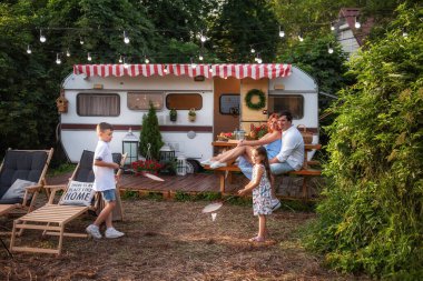 Neşeli erkek ve kız kardeşler karavanda badminton oynuyorlar, mutlu ebeveynler ahşap bankta oturuyorlar. Baba annesine sarılıyor, çocuklar piknikte eğleniyor aile hafta sonları şehir dışında kamp yapıyor.