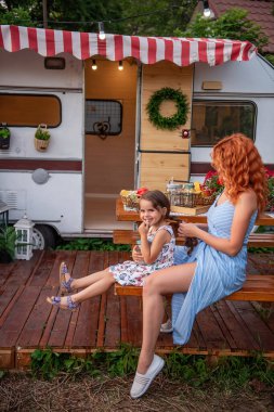 Genç kızıl saçlı anne küçük kızının örgülerini karavan kamyonuyla ahşap bankta örüyor. Annelik bakımı. Kız limonata içiyor. Piknik, şehir dışında ailesiyle kamp yapmak. Çocuklarla Hafta Sonu