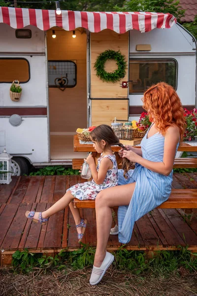 Young red-haired mother braids her little daughter\'s braids on wooden bench by trailer truck. Maternal care. Girl is drinking lemonade. Picnic, camping with family outside city. Weekend with children