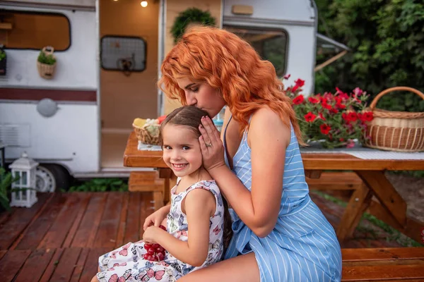 Young red-haired mother braids her little daughter\'s braids on wooden bench by trailer truck. Maternal care. Girl is drinking lemonade. Picnic, camping with family outside city. Weekend with children