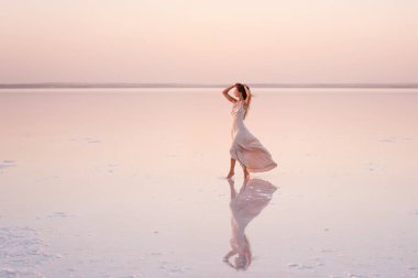 Akşamları havadar, pastel pembeli, pudralı elbisesi olan genç sarışın kadın beyaz kristalize tuz üzerinde yalınayak duruyor. Doğal makyajlı bir kız, saçı gelişiyor. Tuz madenciliği gezisi, gün batımında suda yürümek.