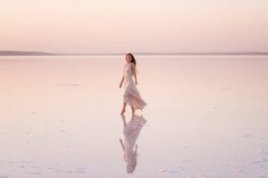 Akşamları havadar, pastel pembeli, pudralı elbisesi olan genç sarışın kadın beyaz kristalize tuz üzerinde yalınayak duruyor. Doğal makyajlı bir kız, saçı gelişiyor. Tuz madenciliği gezisi, gün batımında suda yürümek.