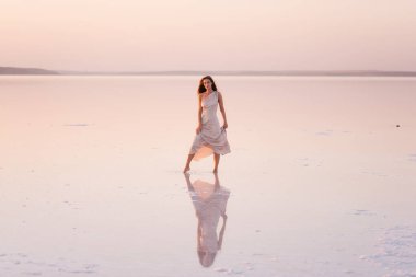 Akşamları havadar, pastel pembeli, pudralı elbisesi olan genç sarışın kadın beyaz kristalize tuz üzerinde yalınayak duruyor. Doğal makyajlı bir kız, saçı gelişiyor. Tuz madenciliği gezisi, gün batımında suda yürümek.