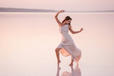 Akşamları havadar, pastel pembeli, pudralı elbisesi olan genç sarışın kadın beyaz kristalize tuz üzerinde yalınayak duruyor. Doğal makyajlı bir kız, saçı gelişiyor. Tuz madenciliği gezisi, gün batımında suda yürümek.