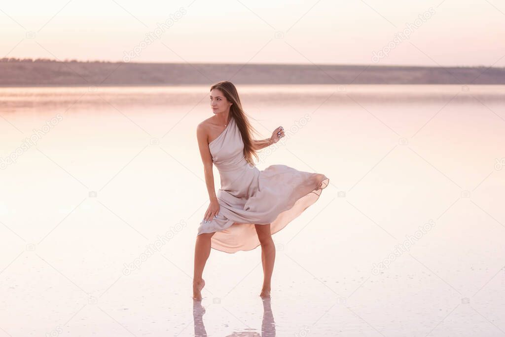 Young blonde woman in an evening airy pastel pink, powdery dress stands barefoot on white crystallized salt. Girl with natural make-up, hair is developing. Salt mining trip, walking on water at sunset