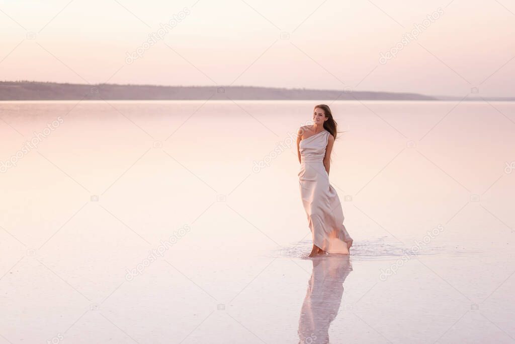 Young blonde woman in an evening airy pastel pink, powdery dress stands barefoot on white crystallized salt. Girl with natural make-up, hair is developing. Salt mining trip, walking on water at sunset