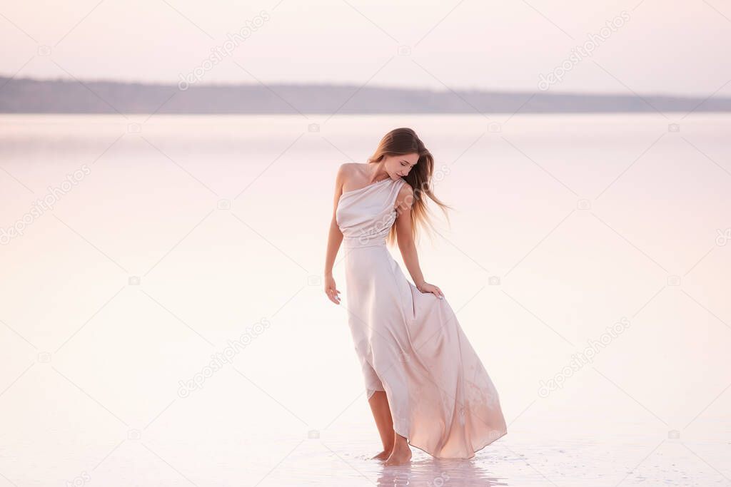 Young blonde woman in an evening airy pastel pink, powdery dress stands barefoot on white crystallized salt. Girl with natural make-up, hair is developing. Salt mining trip, walking on water at sunset