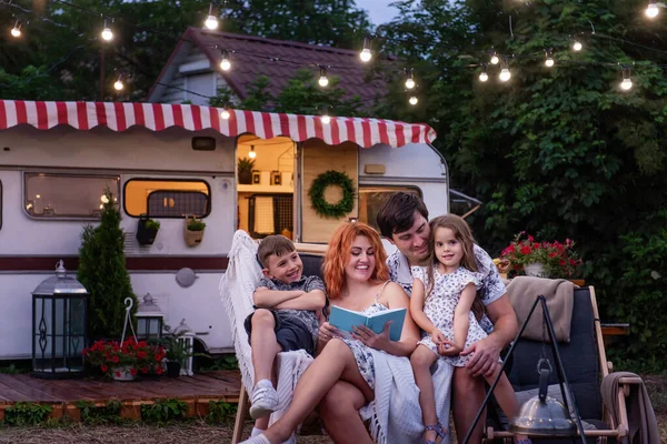 Happy family reads bedtime stories, sitting on sun loungers by the motor at home. Young mother is holding a book in hands, the children and husband are listening nearby. Family weekend countryside