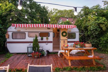 Aile tatilleri için klasik ev karavanı, bütçe seyahati. Ahşap verandada yemek için bankları olan bir masa var. Çimlerin üzerinde güneşlenmek için dinlenme yeri var. İz, lamba çelengiyle süslenmiş