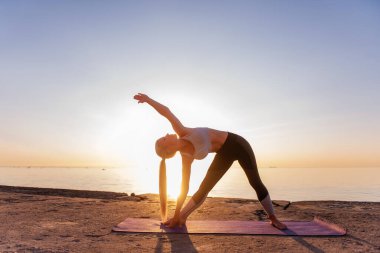Spor kıyafetli genç sarışın kadın yoga yapıyor, güneş doğarken deniz kıyısında egzersiz yapıyor. Kız spora gidiyor, nefes alma egzersizi yapıyor, meditasyon yapıyor, Uzatılmış Üçgen Poz veriyor. Sağlıklı zihin