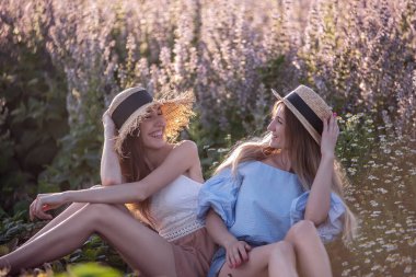 İki kız arkadaş çiçek açan, papatyalı pembe adaçayı tarlasında oturuyorlar. Hasır şapkalı genç kız kardeşler kırsal kesimde şehrin dışında dinleniyorlar. Tarım turunda çiftliğe git.