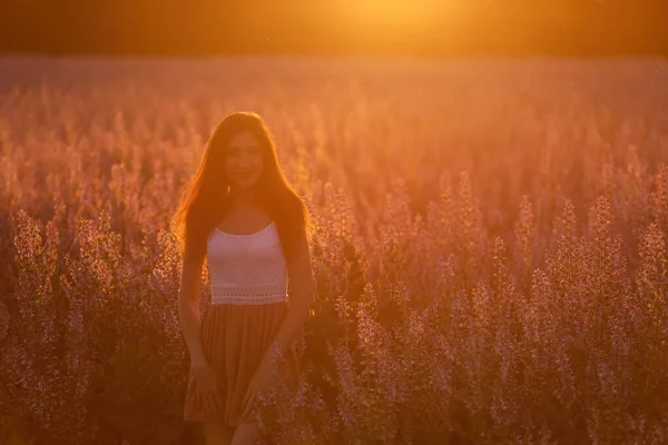 白いTシャツを着た若い女性が ピンクのセージ畑を咲かせている 太陽のオレンジ色の線で肖像画の女の子は 髪の夕日で風になびかせ 花の質感 町を旅行する — ストック写真
