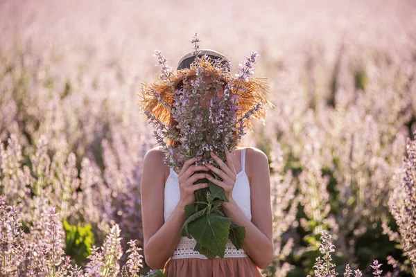 麦藁帽子の若いブロンドの女性は ピンクのセージを咲かせるフィールドの背景に対する距離を見ています 手に花束の花を保持美しい少女のクローズアップ肖像画 農業の食感 — ストック写真