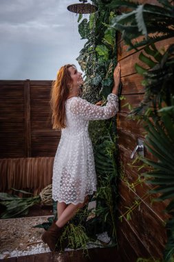 Beyaz elbiseli genç bir kadın, botları duşun altında, tropik yeşil bitkilerle dolu, tahta kahverengi bir duvarın önünde duruyor: canavar, incir ağacı, palmiye ağacı. Gün batımında damlalar bir kızın üzerine düşer.