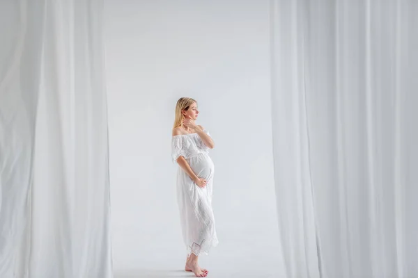 Beyaz elbiseli bir bebek bekleyen hamile bir kadın göbeğine sarılıyor. Havadar kumaşların içinde, minimalist, izole edilmiş bir şekilde duran bir moda kızı. Kolay, mutlu hamilelik, taşıyıcı annelik.