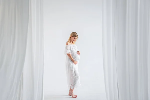 Beyaz elbiseli bir bebek bekleyen hamile bir kadın göbeğine sarılıyor. Havadar kumaşların içinde, minimalist, izole edilmiş bir şekilde duran bir moda kızı. Kolay, mutlu hamilelik, taşıyıcı annelik.