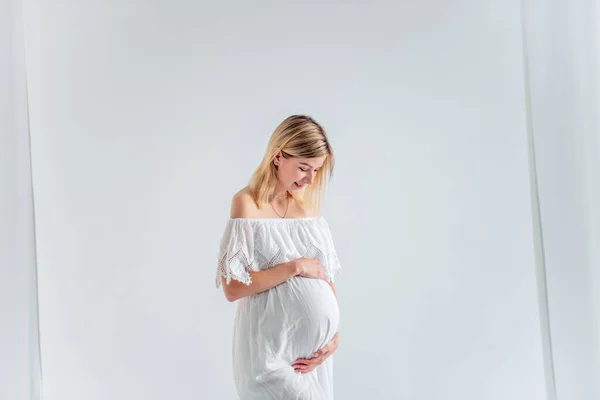 Beyaz elbiseli bir bebek bekleyen hamile bir kadın göbeğine sarılıyor. Havadar kumaşların içinde, minimalist, izole edilmiş bir şekilde duran bir moda kızı. Kolay, mutlu hamilelik, taşıyıcı annelik.