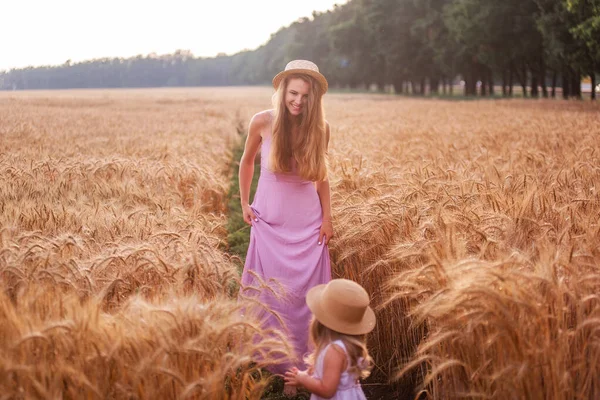 日没時に黄金の小麦畑に沿って歩く母親の手を保持します 若い女性とピンクのドレスとわらの帽子の女の子は ライ麦の中に立って 泥の中に足 田舎のアグロウォーク — ストック写真
