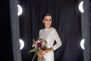 Beyaz şarap rengi peçesiz moda gelini, siyah üzerine açılış elbisesi, öndeki ışıkların arasında kumaş arka plan, elinde buket var. Profesyonel gelin makyajı, dumanlı gözler, şehvetli dudaklar. Saçları topuz gibi olmuş.