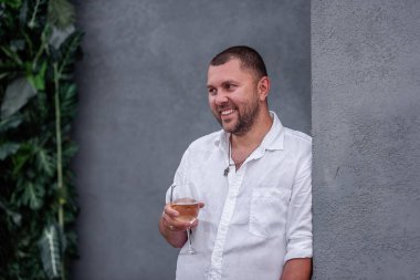 Close-up portrait of a young stylish man holding a glass of white wine, drinking. A guy on a date is wearing a white shirt, blue jeans, standing against a gray wall. The groom laughs at the wedding