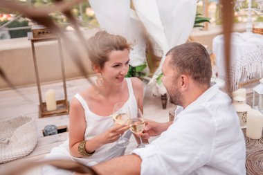 A loving couple sits in a wooden hut in the Moroccan style, drinking white wine. Newlyweds celebrate their anniversary. Young woman laughs, hugs bearded man on open luxury roof terrace with sea view.
